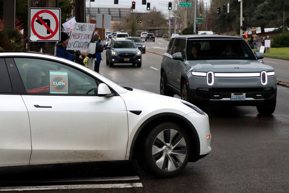 A Tesla Model Y with an anti-Elon Musk sign in the window, with a Rivian R1S and protestors