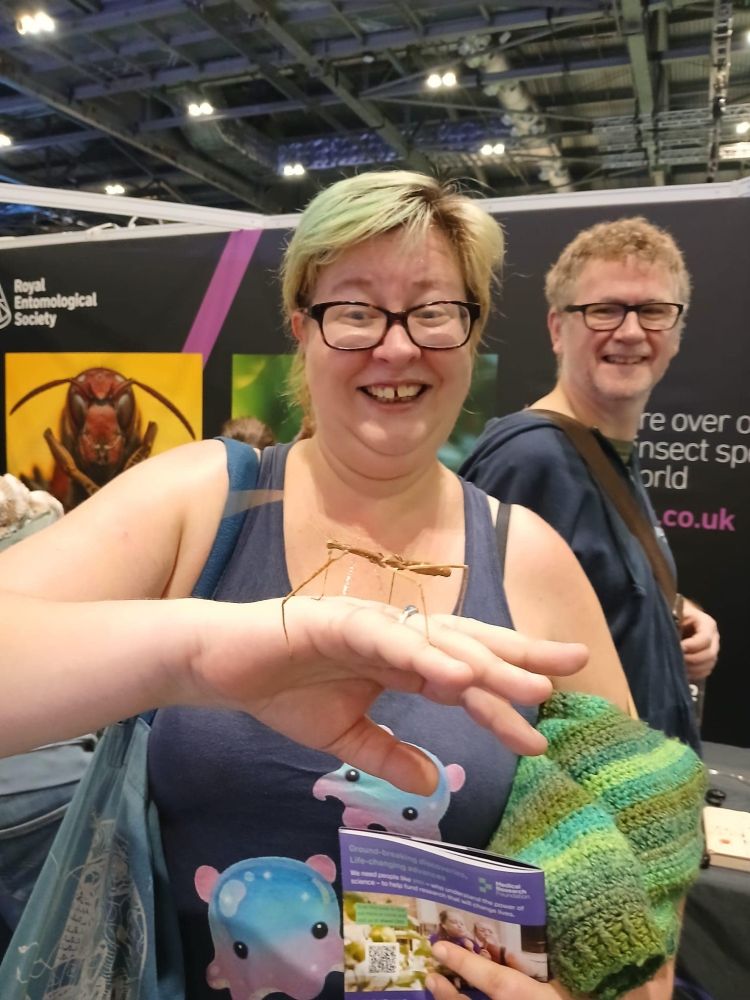 A photo of me (white woman, short bleached blonde hair, glasses). My hand is outstretched, palm down, and on the back of my hand is perched a stick insect that is as long as my palm. I look *delighted* 