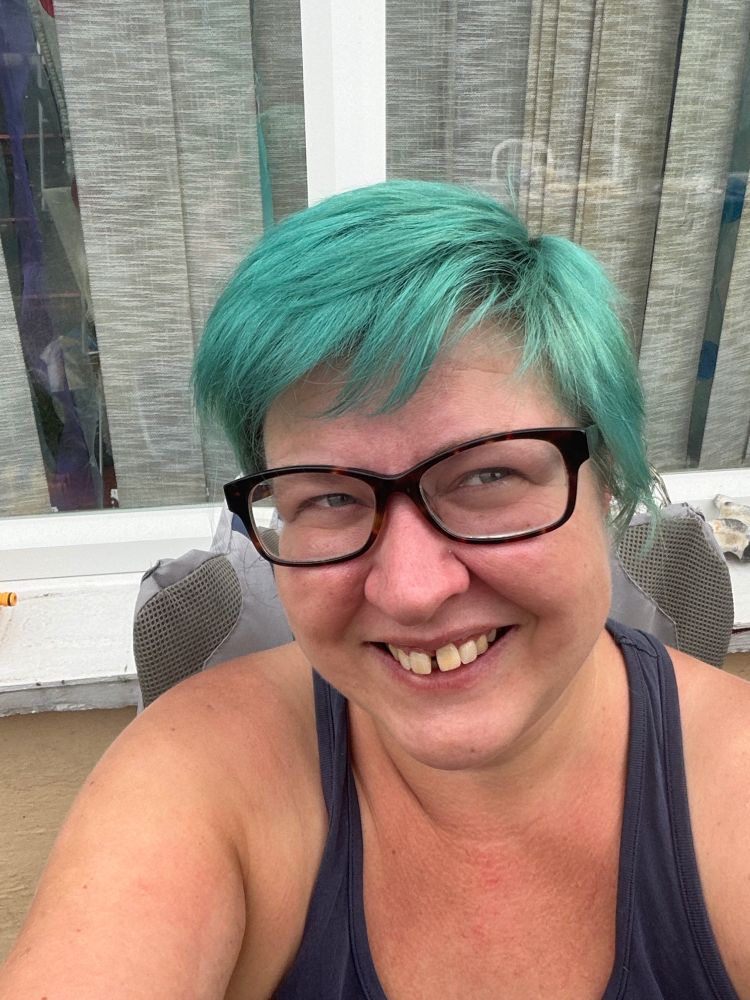 A selfie of a white woman wearing chunky plastic-framed glasses, with short hair in a side parting. The hair is a blue/green mermaid colour. The woman (me) is looking up towards the top right of the frame and is smiling. 