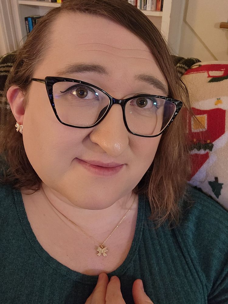 A white woman in her 40s with shoulder-length brown hair, wearing black cat-eye glasses, an emerald green sweater, and matching gold bowtie earrings and necklace