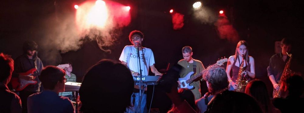 6 band members against a dark moody nightclub background, dry ice emerging from the main stage light by the singer and keyboardist, to the right of him the bass and 2 saxophones, to the left drummer and guitarist.  