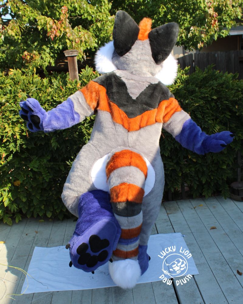 A grey, black, orange and purple red panda fursuit facing away from the camera showing off its toe beans.