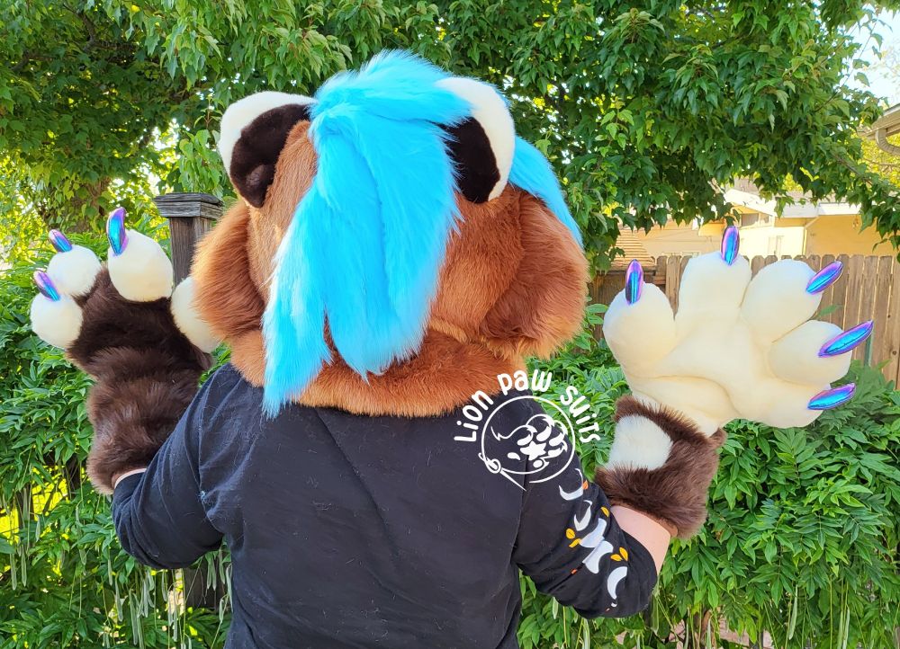 A brown and cream mustelid fursuit with turquoise hair and holographic claws facing away from the camera to show off their hair.