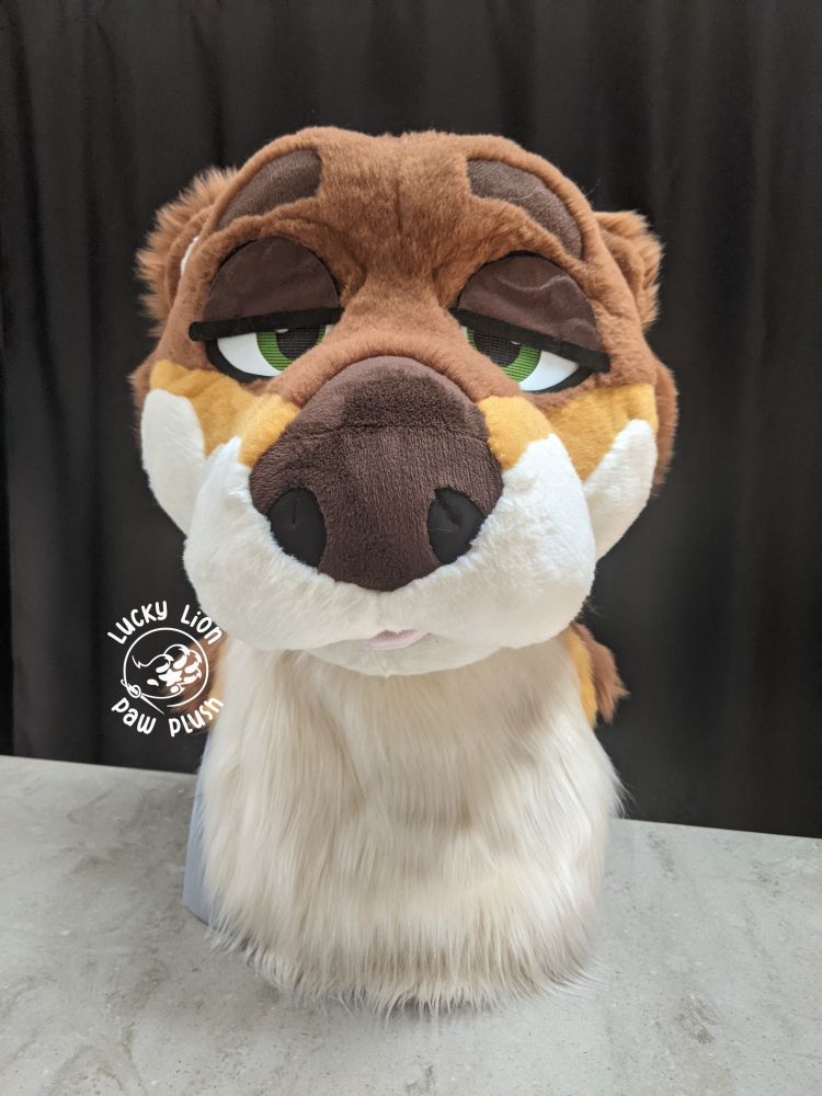 A brown and cream otter fursuit head with gold accents and green eyes showing off 'sleepy' eyelids. 