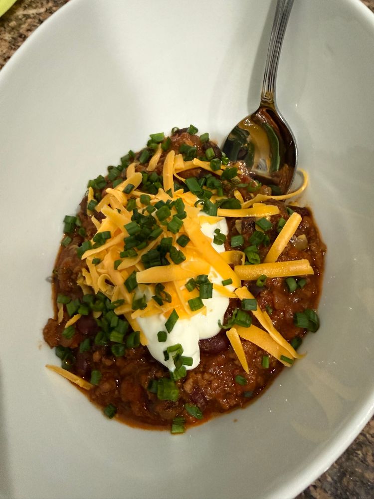 A white bowl with red chilli topped with sour cream, cheddar cheese and chives. There is a spoon in there as well