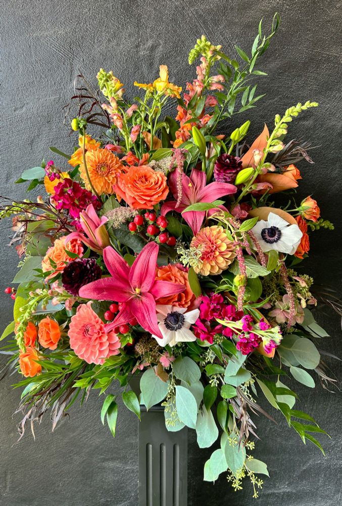 A large floral design including dark pink lilies orange, pink,  and yellow dahlias, magenta stock, orange roses, orange spray roses, mauve lace hydrangea, purple ranunculus, pink snapdragons, white anemones, red hypericum berries, yellow butterfly ranunculus, burgundy lace flower, and red hanging amaranth. The greenery consists of italian ruscus, agonis, weeping podocarpus, seeded and silver dollar eucalyptus