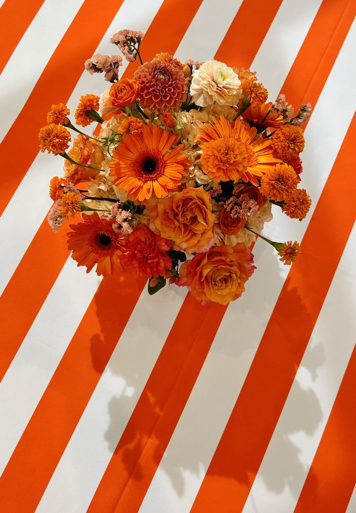 An overhead view of a floral arrangement with various shades of orange flowers. The arrangement is sitting on an orange and white striped tablecloth, and casting a shadow from the sun onto the tablecloth.