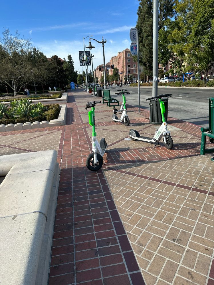 Photo of brick sidewalk next to street, with 3 electric scooters that are left blocking the path.