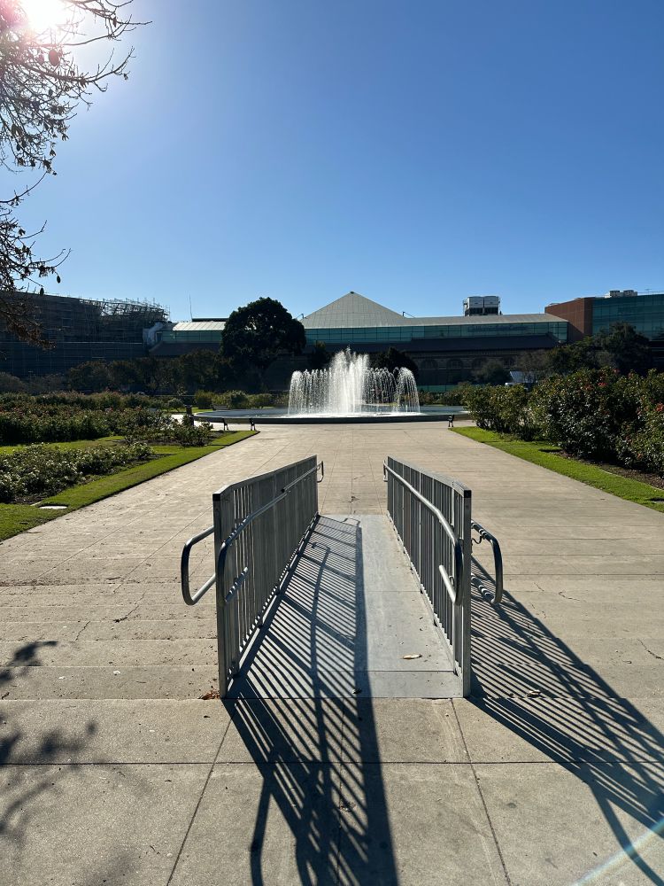 Photo of a metal wheelchair ramp that has been added to path of concrete steps. A water fountain in the background and building with pyramid roof. Rose gardens on the sides. Clear blue sky.