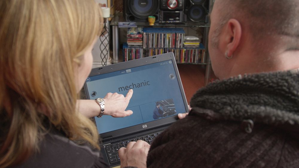 Photo of a speech and language therapist helping and pointing to words on a computer screen for a patient who is looking at the screen.