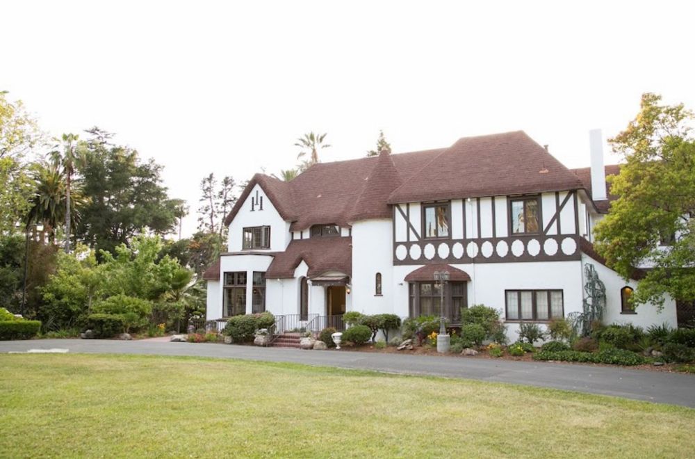 Photo of tudor style castle-cottage house, brown roof, brown window trims, small tower spire. Green grass and driveway out front. Trees and bushes around the front and sides. Pathway to the back.