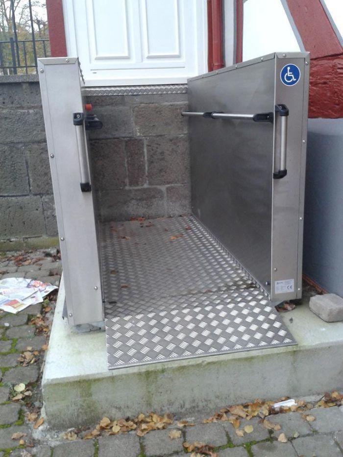 Photo of metal wheelchair lift, outside of backdoor, on cobblestone sidewalk with leaves, next to stone wall, with white backdoor above.