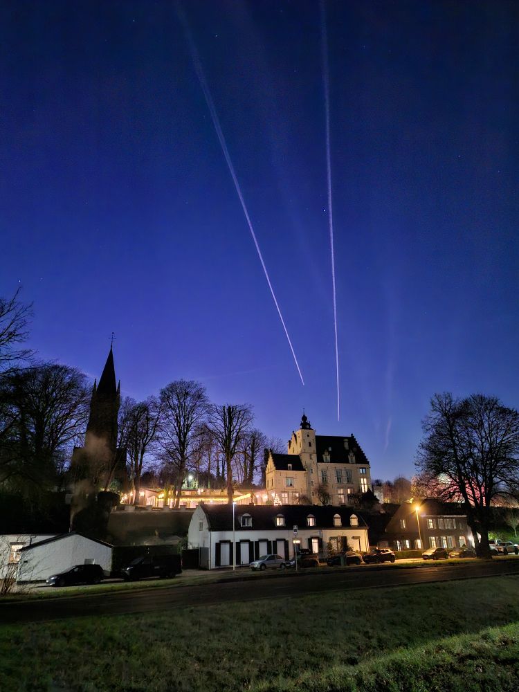 Airplanes cut through the clear evening sky above a brightly lit mansion. A gothic church is silhouetted in the backdrop