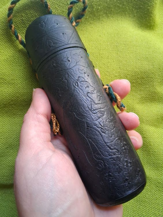 A cylindrical black leather case, held in a white hand, on a bug green background. The case is incised with floral decorations and an upright standing bear.