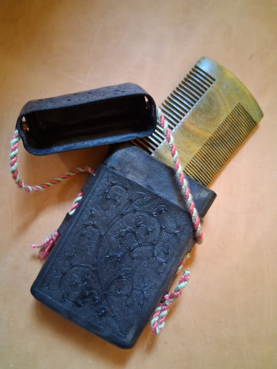 A black leather case etched with vines lies open on a natural leather background. A green sandalwood comb is partially inside it.