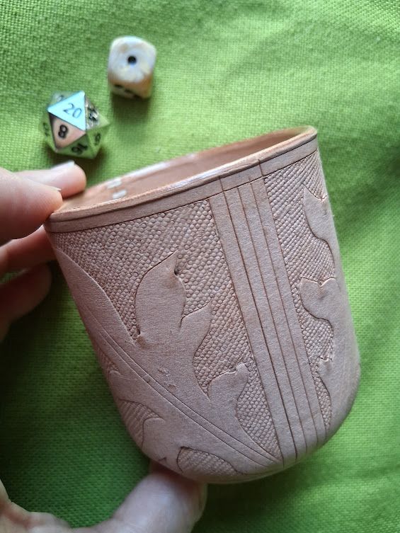 A handheld cup made of natural leather, with a large leaf design and small circle background. Two dice are out of focus in the upper left corner, to demonstrate the function.