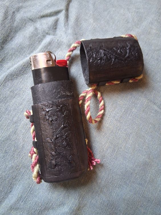 A black leather case with a medieval vine motif carved into it. It has a pink and yellow cord running through loops on the side. It is on a gray cloth background. Inside the case is a standard black plastic bic lighter.