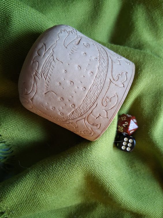 A leather cup on a green background, with two dice spilling out. The cup is decorated with a cute medieval frog.