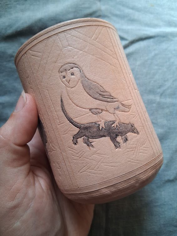 A natural leather dice cup held in a pale hand, on a blue-gray background. The cup is decorated with a barn owl grasping a rat.