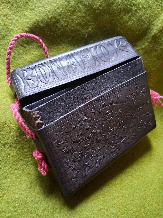 A black leather card case on a green felt background. The visible lettering reads "BON AMOR".