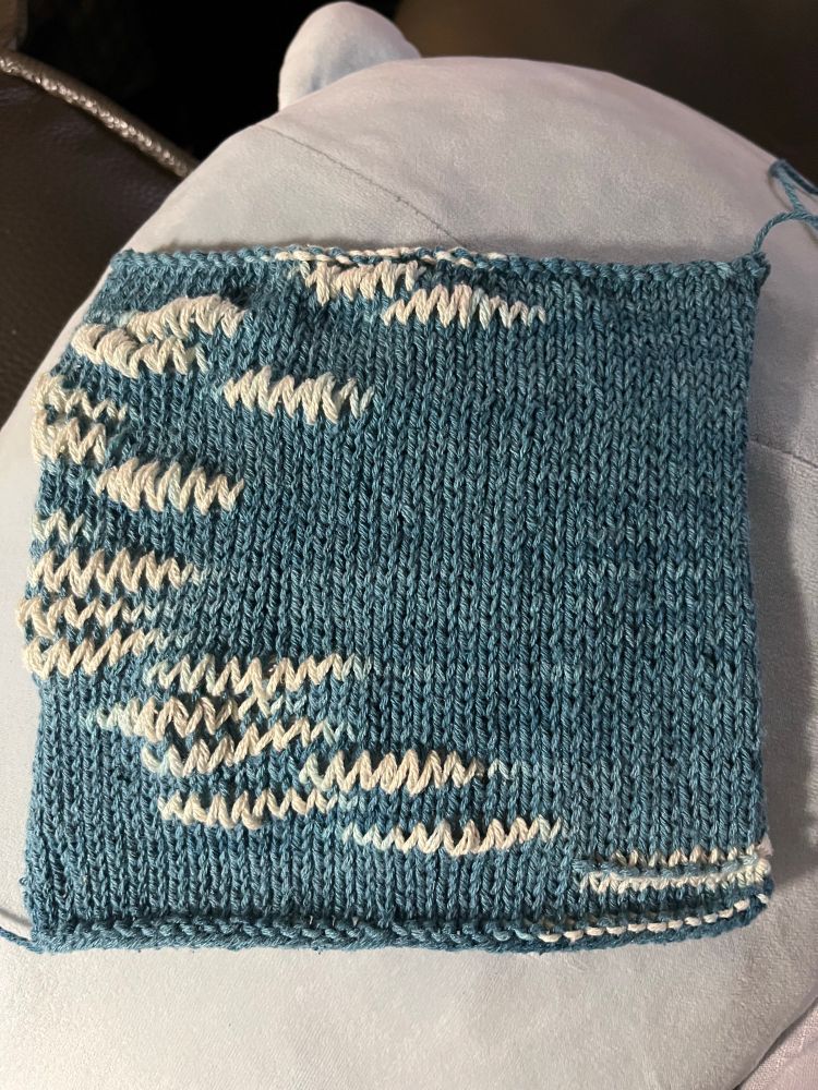 A square of knitted medium blue fabric with little sections of taller stitches in the natural color. They look like little squiggles.
