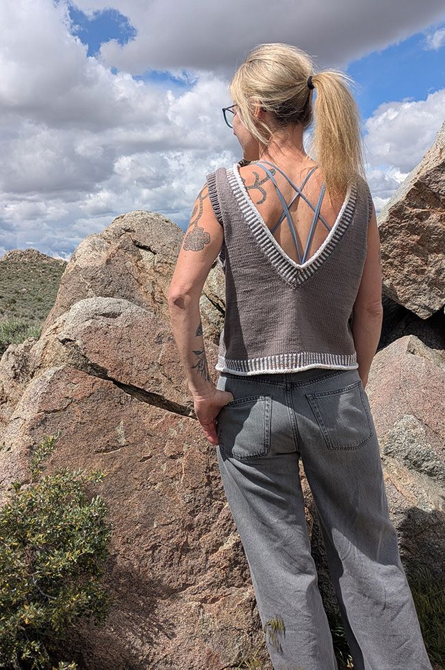 Back view of a woman with long blond hair in a ponytail. She is wearing a waist-length sleeveless sweater with a very deep V back in a medium taupe color. The sweater has corrugated ribbing trim at the hem and neck. 