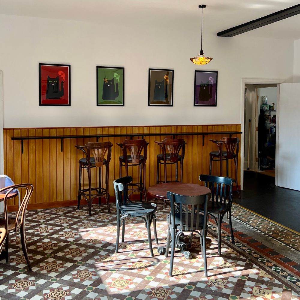 A cosy café interior featuring a patterned tile floor, a mix of dark wooden chairs and tables, and a wooden half-wall with high stools along a bar ledge. Four framed cat illustrations with colourful backgrounds (red, green, brown, and purple) hang on the white wall above the bar.