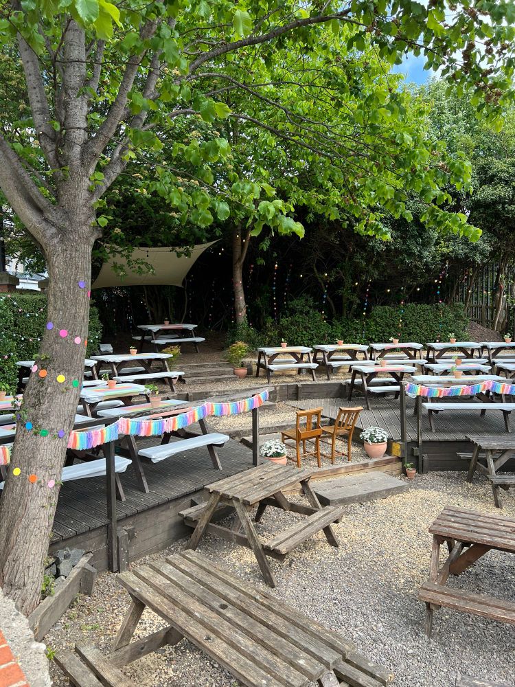 A vibrant outdoor seating area in The Free Trade Inn’s beer garden with multiple wooden picnic benches arranged on different levels. The space is decorated with colourful paper garlands and string lights wrapped around trees and hanging from branches. There are two small wooden chairs placed on a circular gravel area, surrounded by potted flowers. The area is shaded by lush green trees and a large white canopy in the back provides additional cover.