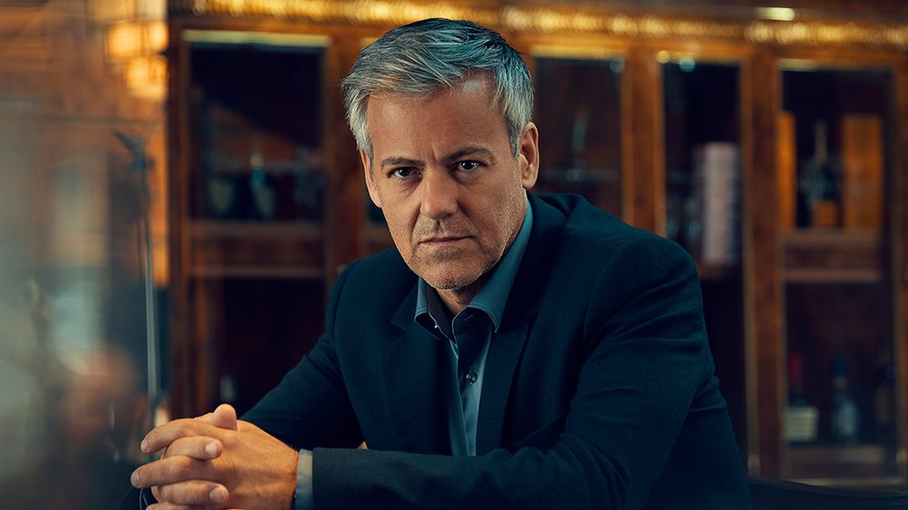 A picture of Rupert Graves as Gabriel Hirsch in Series 3 of "Riviera" in 2020. He is sitting at a bar with his arms resting on it and his hands folded, looking directly at the camera as if it is the bartender. There are wooden cabinets full of expensive bottles behind him.