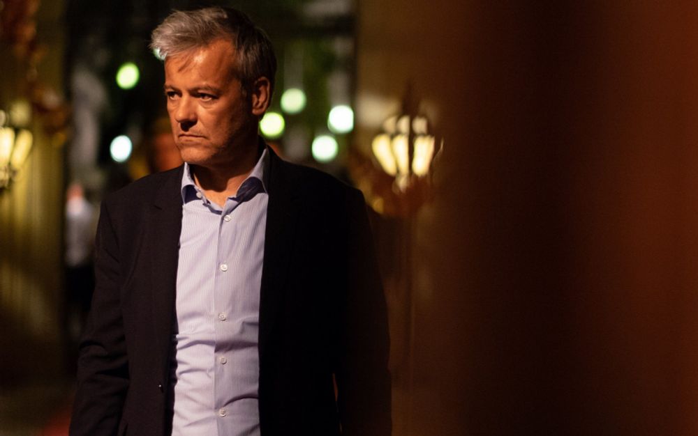 Rupert Graves as Gabriel Hirsch in Riviera. He is wearing a dark suit and white shirt, standng by a canal in Venice at nighttime, looking very serious.