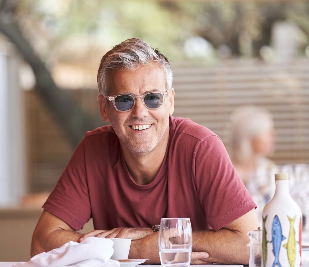 Rupert Graves as Gabriel Hirsch in Riviera. He is wearing a brick red t-shirt and sunglasses, having breakfast in a restaurant and smiling at someone off to the right.