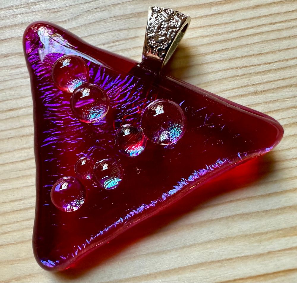 A pinkish-red triangle pendant with glass dots that have a slight metallic blue sheen to them. It has a silver bail and is laying on a light wooden table.