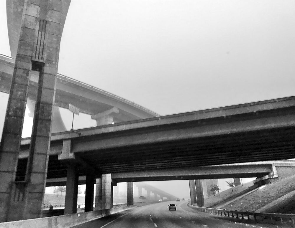 Black and white photo of a highway road passing under an overpass. To the left are two more overpasses even higher up. 