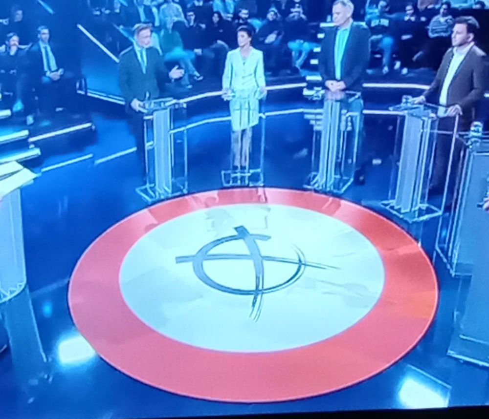Foto von TV-Gerät mit Talkrunde ZDF, "Schlagabtausch". Die Talkgäste stehen im Kreis. In der Mitte ist ein roter Kreis auf dem Boden abgebildet, im inneren dessen eine weiße Fläche. In der Mitte der weißen Fläche ein handschriftliches, schwarzes Kreuz in einem gezeichneten schwarzen Kreis (der schwarze Kreis und das Wählerkreuz sehen aus wie ein Hakenkreuz). 