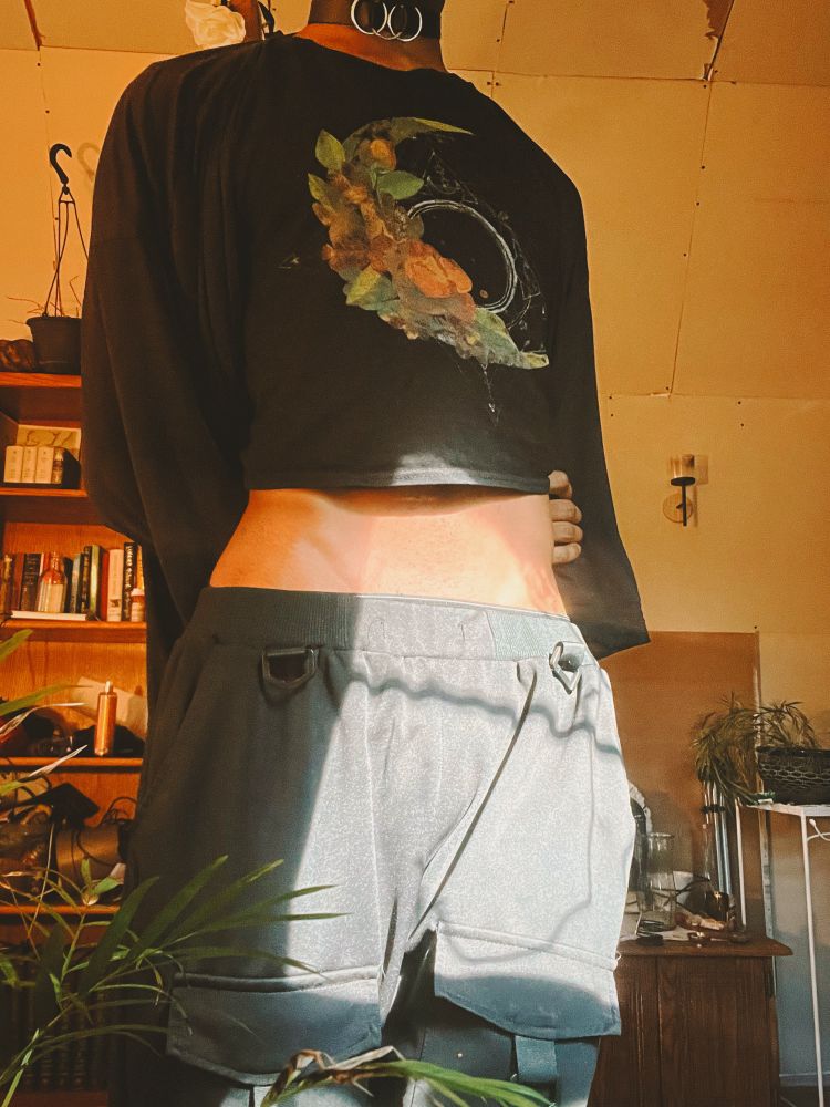Darling stands dressed in a black crop top, sweat and pants. The sunlight shines on their exposed hips 