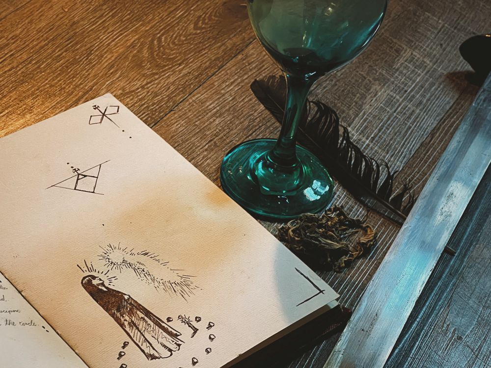 A photo featuring the partial page of darling’s spell book. The page features and illustration of an incantation to speak to a spirit and various signal. Beside the book or feather, a few pieces of mugwort, the blade of a sword, and a chalice.