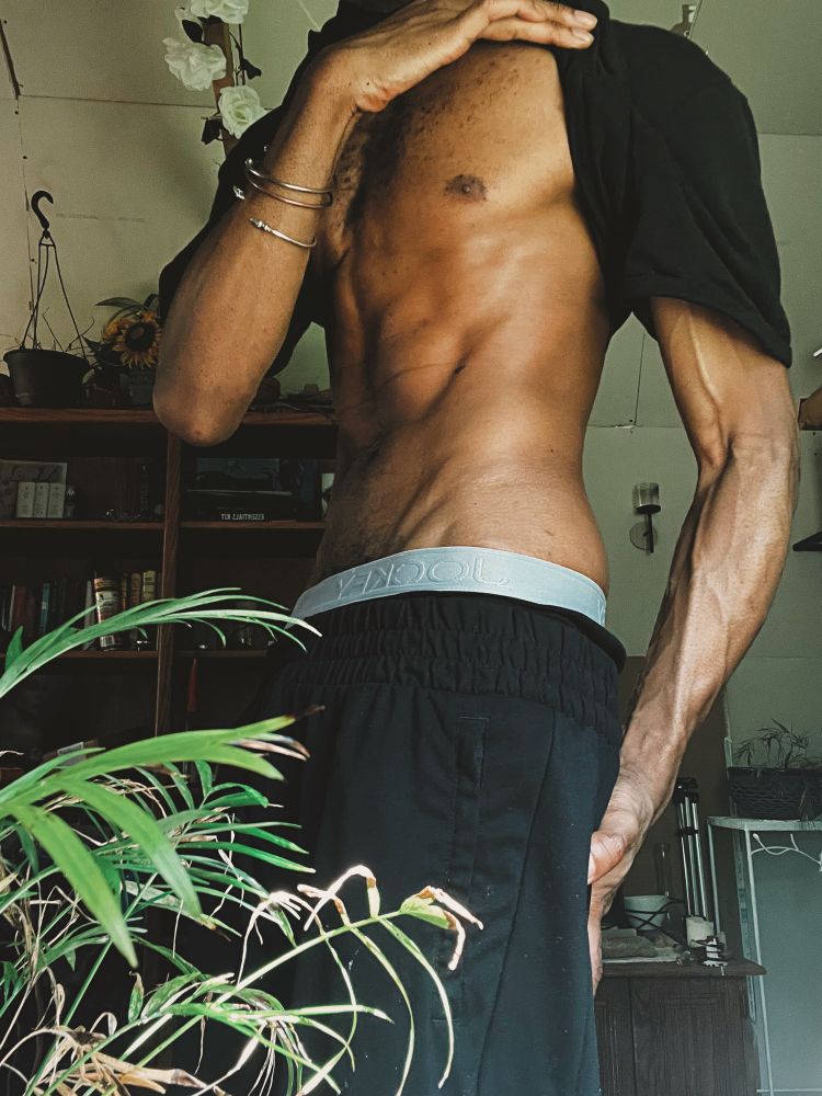 Darling lifts the shirt of their all-black attire to show off their toned torso from the side while the veins of their arm are quite prominent 