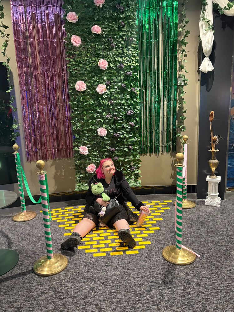 Me (a white gal with pink hair wearing a black outfit) sitting bc on a a “yellow brick road” floor with my little green build a bear frog (who is dressed as a witch). Behind me is a pink and green wall with tinsel for the wicked movie 