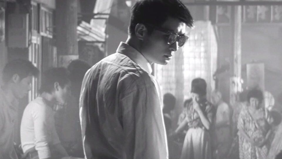 Young man in a crowded, smoky bar looking too cool for school. He's wearing sunglasses and a white shirt