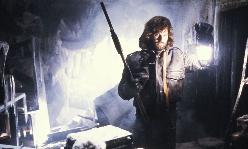 Kurt Russell holding a shotgun in one hand and a hurricane lamp in the other. Standing in an ice covered hut