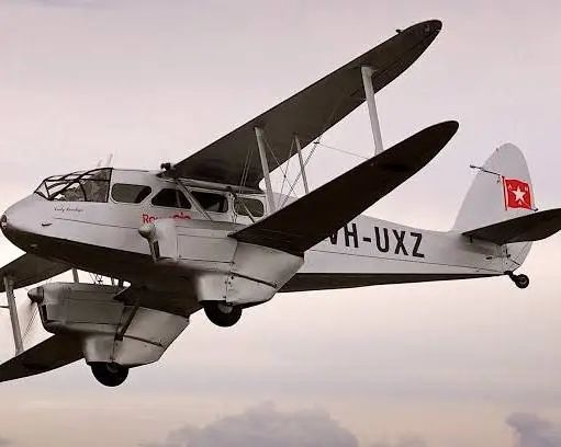 A DeHavilland Dragon Rapide biplane airliner with its distinctive swept-lobe tail flies through a grey sky.