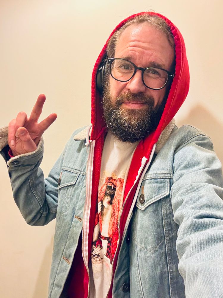 A person with a full beard and glasses is taking a casual selfie indoors. They’re wearing a red hoodie with the hood up under a light denim jacket. Their T-shirt features a bold graphic of a figure with a bear’s head behind them. They’re holding up a peace sign with a relaxed expression.
