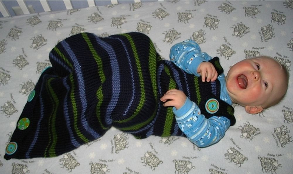 A laughing baby in his crib wearing a blue and green striped handknit sleep sack. 
