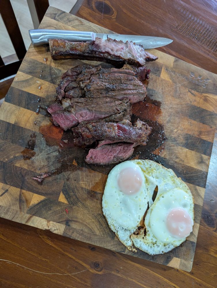 A wooden cutting board features a fully cooked King ribeye steak, sliced into thick pieces to reveal its tender, medium-rare interior. Beside the steak, two perfectly fried eggs with golden, runny yolks add a vibrant contrast. A large knife rests atop the steak slices, emphasizing the rustic presentation. The cutting board showcases a geometric pattern, and beneath it is a wooden table. 