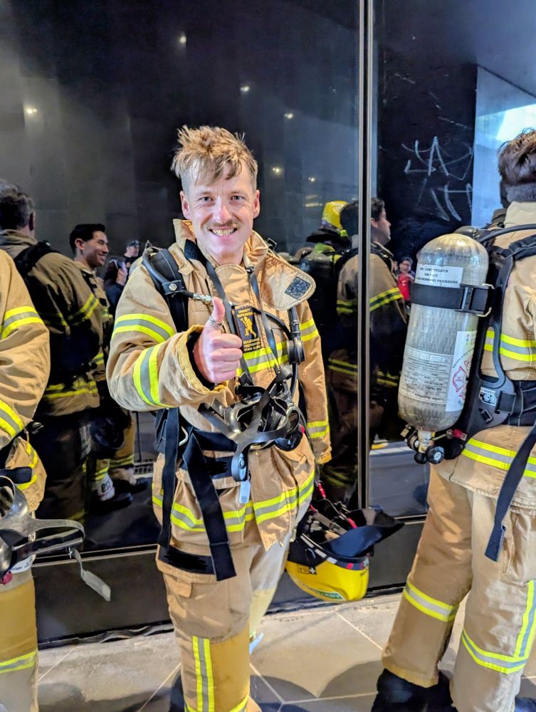 A firefighter in full gear stands indoors, smiling and giving a thumbs-up. The gear includes a tan structural firefighting suit with reflective yellow and silver stripes, and an oxygen tank strapped to the back. The firefighter holds a helmet in the left hand. Behind them, other firefighters in similar gear are visible, some partially obscured. 
