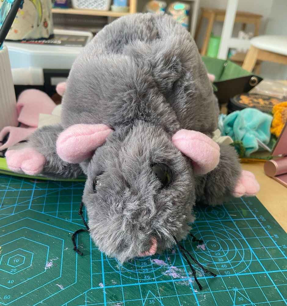 big fat fluffy grey plush rat with pink ears and feet lying on a cluttered table