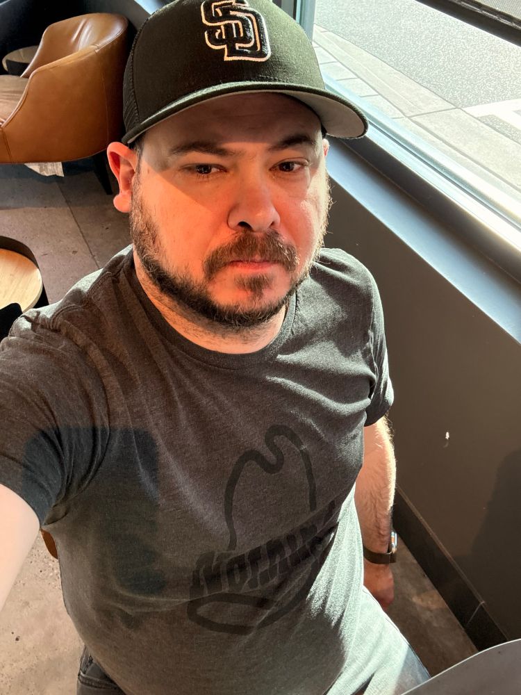 Lanky white guy taking a selfie in a seated position from a high, downward angle. He is wearing a black San Diego Padres batting practice low-profile 5950 fitted cap with “SD” in white outline. His black shirt says “Nothing” within an Arby’s hat in a deeper shade of black. He is wearing black jeans (with a flexible waist, thank goodness).