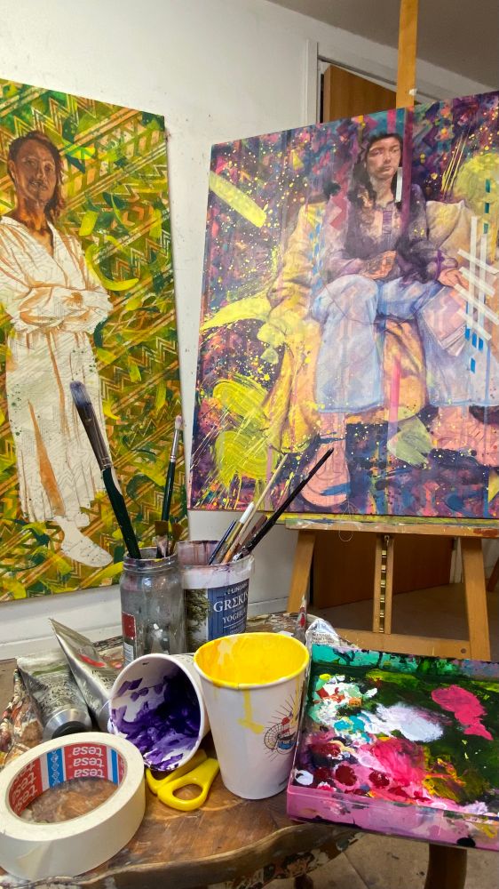 A painter’s paraphernalia in the foreground. In the background 2 large and unfinished portraits of women on heavily patterned and colourful backgrounds