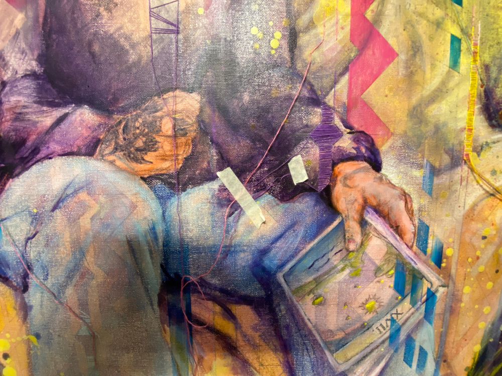 Close up of a portrait in progress. Focus is on the unfinished hands of the subject. The right hand is resting on a knee lightly closed. The left hand is hanging slightly off the knee holding a book by it’s spine 