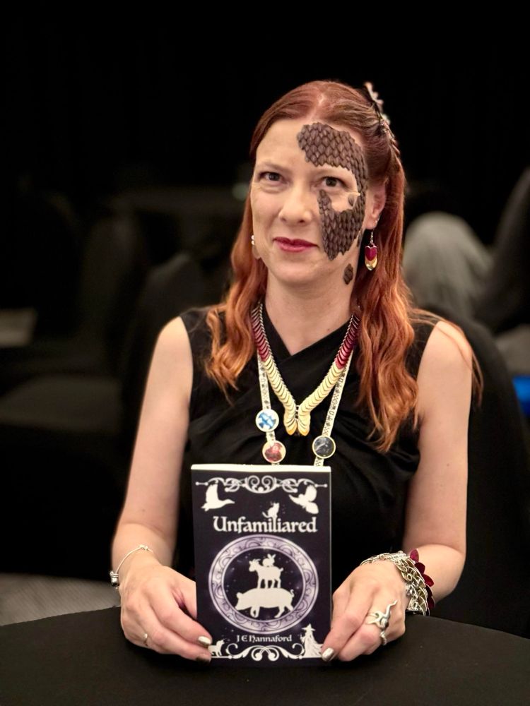 J E Hannaford with scales on her face sat holding a copy of her novella, Unfamiliared 